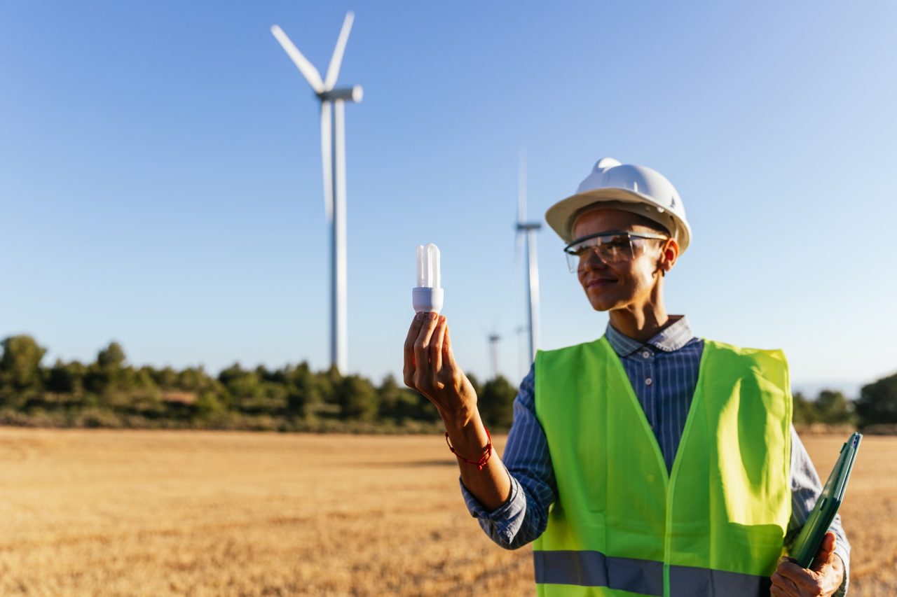 Ingeniero eléctrico sosteniendo una bombilla de bajo consumo sobre un fondo desenfocado de un molino de viento